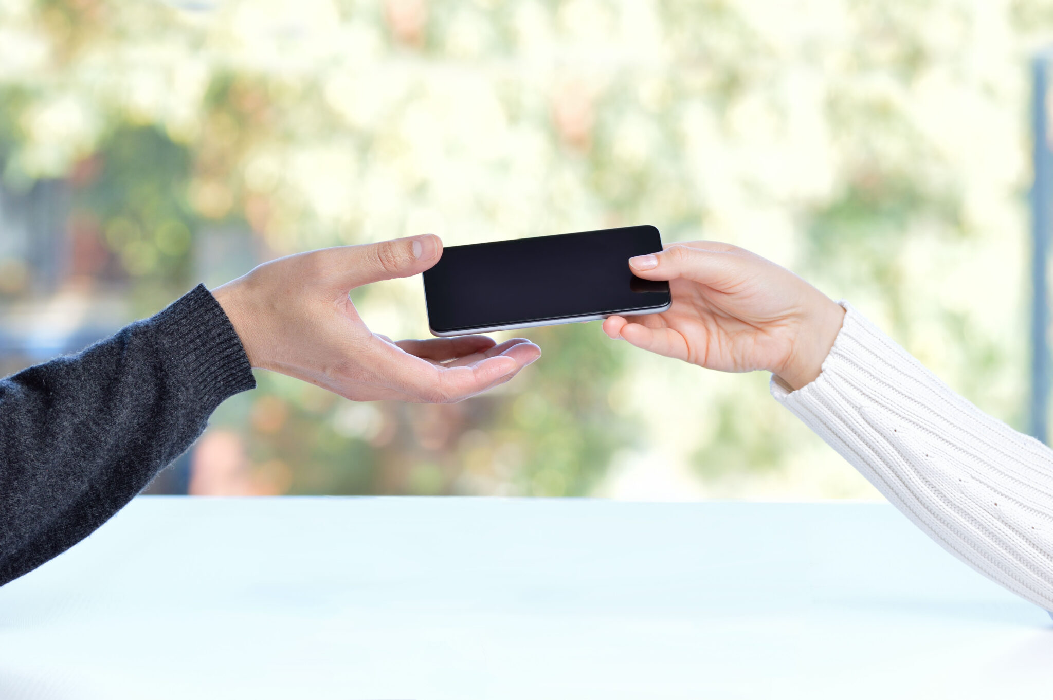 Zwei Hände übergeben sich ein Smartphone. Die linke Hand trägt einen grauen, die rechte einen weißen Pullover. Der Hintergrund ist unscharf und zeigt eine grüne Landschaft.