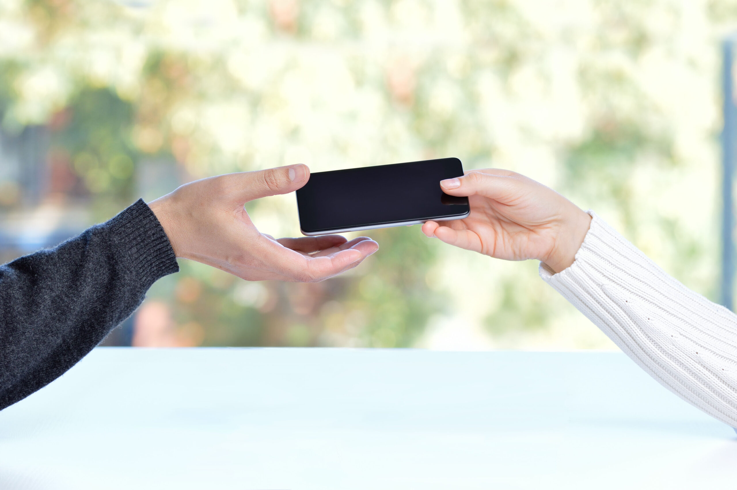 Zwei Hände übergeben sich ein Smartphone. Die linke Hand trägt einen grauen, die rechte einen weißen Pullover. Der Hintergrund ist unscharf und zeigt eine grüne Landschaft.