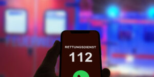 Ein Smartphone-Bildschirm zeigt die Nummer 112 mit der Aufschrift 'Rettungsdienst' und einem grünen Anrufsymbol. Im Hintergrund ist ein unscharfes Rettungsfahrzeug mit blinkenden Lichtern zu sehen.