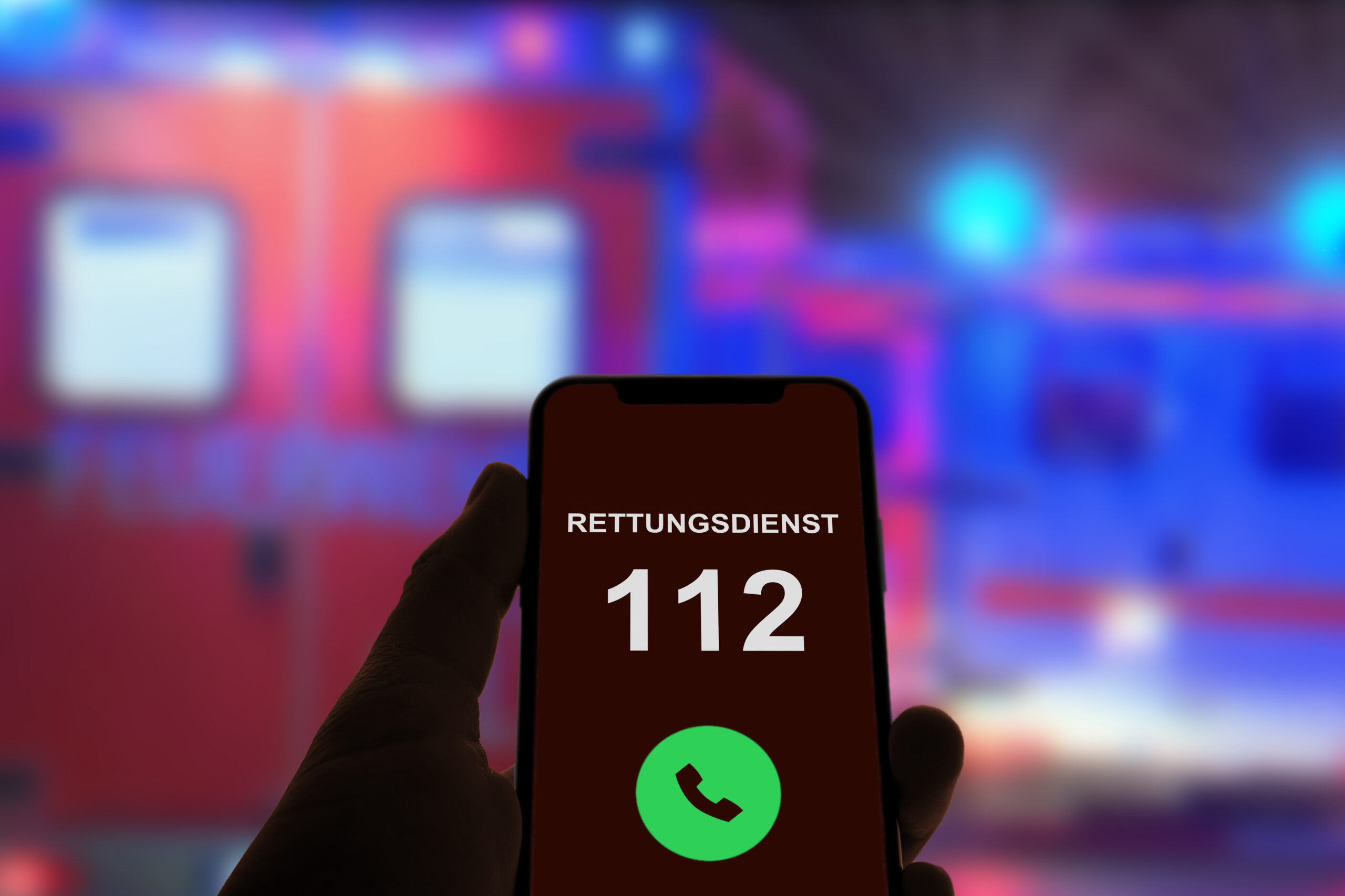 Ein Smartphone-Bildschirm zeigt die Nummer 112 mit der Aufschrift 'Rettungsdienst' und einem grünen Anrufsymbol. Im Hintergrund ist ein unscharfes Rettungsfahrzeug mit blinkenden Lichtern zu sehen.