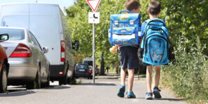 Zwei Jungen mit bunten Schulranzen gehen auf einem Gehweg entlang, neben ihnen parkende Autos und Bäume. Im Hintergrund ist ein Vorfahrtsschild zu sehen, während sie auf dem Weg zur Schule sind.