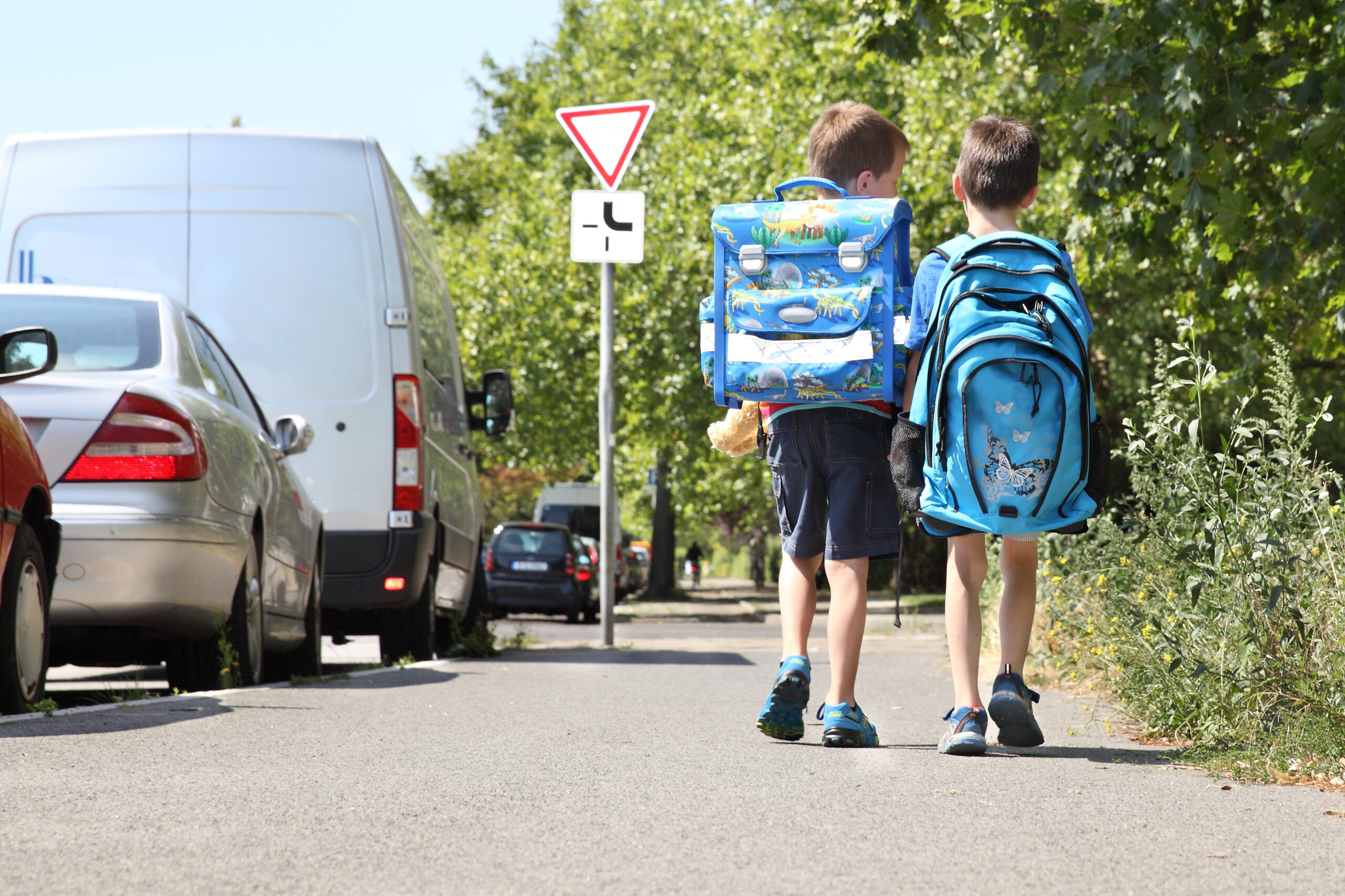 Zwei Jungen mit bunten Schulranzen gehen auf einem Gehweg entlang, neben ihnen parkende Autos und Bäume. Im Hintergrund ist ein Vorfahrtsschild zu sehen, während sie auf dem Weg zur Schule sind.